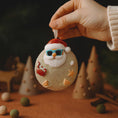 Load image into Gallery viewer, Hand holding a decorative ornament with a Santa Claus face, surrounded by small trees and items on a dark background.