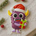 Load image into Gallery viewer, Purple monster with a Santa hat holding a gift on a snowy background