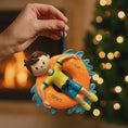 Load image into Gallery viewer, Decorative ornament of a child on an inner tube with a blurred Christmas tree and fireplace in the background.