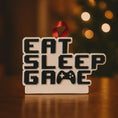 Load image into Gallery viewer, Personalized Eat Sleep Game Christmas ornament with a raised 3D front featuring bold block-style lettering and a detailed game controller icon. The front design is fully three dimensional, creating depth and texture, while the back is flat 2D for easy hanging and durability. White base with black raised letters and subtle colored accents. Includes hand-added personalization with name and year or family name. Gaming-themed holiday ornament displayed on snowy festive background with warm lights, designed for