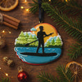 Load image into Gallery viewer, Decorative ornament of a person paddleboarding on a lake with trees and sun, placed on a wooden surface with Christmas decorations.