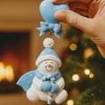Load image into Gallery viewer, Decorative snowman ornament with blue hat and scarf held by a hand against a festive background.