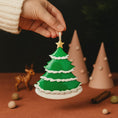 Load image into Gallery viewer, Hand holding a green Christmas tree ornament with a star on top, surrounded by festive decorations.