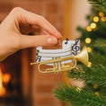 Load image into Gallery viewer, Personalized music ornament featuring a white musical staff design with black notes and a gold trumpet detail. Includes free handwritten personalization with one name and year written on the sheet music. Shown hanging with a ribbon in a warm festive holiday setting with soft lights instruments and seasonal decor. Designed with a 3D raised front and flat back perfect for Christmas tree display and gifts for musicians music lovers and band members.