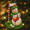 Load image into Gallery viewer, Snowman Christmas ornament with golf club and flag, against a blurred Christmas tree background.