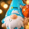 Load image into Gallery viewer, Personalized basketball gnome Christmas ornament featuring a cute blue gnome character holding a basketball, customizable name area on the hat and customizable year area on the base, glossy finish with raised 3D front details and flat 2D back, white hanging ribbon, displayed on a snowy holiday background with pine branches and festive decorations.