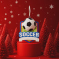 Load image into Gallery viewer, Personalized soccer Christmas ornament featuring a classic black and white soccer ball on a shield style background, customizable name area on the front banner and customizable year area on the soccer ball, glossy finish with raised 3D front details and flat 2D back, white hanging ribbon, displayed on a snowy soccer field themed holiday background with pine branches pinecones and festive decorations.