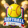 Load image into Gallery viewer, Personalized softball Christmas ornament featuring a yellow softball with stitched details and crossed bats design on a shield style background, customizable name area on the front banner and customizable year area on the softball, glossy finish with raised 3D front details and flat 2D back, white hanging ribbon, displayed on a snowy softball field themed holiday background with pine branches and festive decorations.