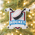 Load image into Gallery viewer, Personalized hockey Christmas ornament featuring a hockey puck and crossed hockey sticks design, customizable name area on the front banner and customizable year area on the puck, glossy finish with raised 3D front details and flat 2D back, white hanging ribbon, displayed on a snowy ice rink themed holiday background with hockey accessories and festive winter decorations.