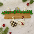 Load image into Gallery viewer, Decorative wall hanging with mittens, candles, and holly leaves on a textured surface.