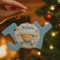 Load image into Gallery viewer, Decorative ornament with a baby face and blue letters spelling 'JOY' against a Christmas tree background.