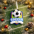 Load image into Gallery viewer, Personalized soccer Christmas ornament featuring a classic black and white soccer ball on a shield style background, customizable name area on the front banner and customizable year area on the soccer ball, glossy finish with raised 3D front details and flat 2D back, white hanging ribbon, displayed on a snowy soccer field themed holiday background with pine branches pinecones and festive decorations.