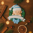 Load image into Gallery viewer, Personalized Baby Boy Christmas Ornament featuring a sweet child peeking from a festive green wreath with blue accents and polka dot bow. This ornament has a raised 3D front design with detailed holly leaves, textured elements, and dimensional lettering. The name is placed on the hat and the year on the banner. A meaningful keepsake for babyâs first Christmas, newborn gift, or holiday memory decoration for the Christmas tree.