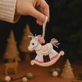Load image into Gallery viewer, Hand holding a pink rocking horse ornament with a blurred Christmas scene in the background.
