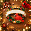 Load image into Gallery viewer, Personalized cardinal wreath ornament featuring a vibrant red cardinal bird surrounded by green holly leaves and red berries. Designed with a smooth glossy finish and a raised 3D front with a flat back for easy hanging. Includes free handwritten personalization with one name and year displayed on the banner at the top. Styled in a warm festive holiday setting with soft Christmas lights candy details and seasonal decor. A meaningful Christmas tree ornament symbolizing remembrance love and connection perfect