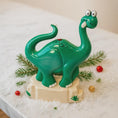 Load image into Gallery viewer, Green dinosaur-shaped night light on a snowy surface with Christmas decorations.