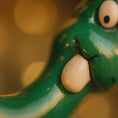 Load image into Gallery viewer, Close-up of a green resi ornament dinosaur with a blurred yellow background