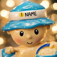 Load image into Gallery viewer, Personalized Beach Baby Christmas Ornament featuring a cheerful toddler sitting on the sand with a beach hat, float, bucket, and shovel. This detailed 3D front design includes raised elements, bright summer colors, and textured sand base for added depth. The name is placed on the hat and the year on the sandy base. A perfect keepsake for baby’s first beach trip or summer memory, ideal for Christmas tree decor or a thoughtful holiday gift for new parents.