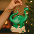 Load image into Gallery viewer, Green dinosaur ornament being held against a blurred Christmas tree background