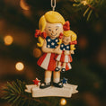 Load image into Gallery viewer, Decorative Christmas ornament of a girl holding a smaller figure, both wearing American flag-themed outfits, hanging on a tree.