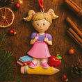 Load image into Gallery viewer, Decorative ornament of a girl with a backpack on a pencil, surrounded by Christmas decorations on a wooden surface.