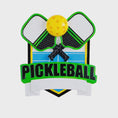 Load image into Gallery viewer, Personalized pickleball Christmas ornament featuring crossed pickleball paddles and a yellow pickleball on a shield style background, customizable name area on the front banner and customizable year area on the ball, glossy finish with raised 3D front details and flat 2D back, white hanging ribbon, displayed on a snowy pickleball court themed holiday background with pine branches and festive decorations.