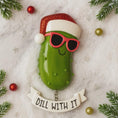 Load image into Gallery viewer, Decorative pickle ornament with sunglasses and Santa hat on a festive background