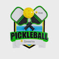Load image into Gallery viewer, Personalized pickleball Christmas ornament featuring crossed pickleball paddles and a yellow pickleball on a shield style background, customizable name area on the front banner and customizable year area on the ball, glossy finish with raised 3D front details and flat 2D back, white hanging ribbon, displayed on a snowy pickleball court themed holiday background with pine branches and festive decorations.