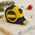 Load image into Gallery viewer, Yellow and black tape measure on a decorative surface with Christmas elements.