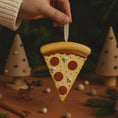Load image into Gallery viewer, Pizza slice ornament being held above a festive scene with Christmas trees and decorations.
