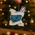 Load image into Gallery viewer, A cozy knitting yarn ornament featuring a detailed blue ball of yarn wrapped with flowing ribbon and raised knitting needles. The ornament has a sculpted 3D front with realistic yarn texture and smooth glossy finish, while the back is flat and 2D for easy hanging. Personalized with a handwritten name and year, this resin ornament is displayed in a warm festive setting with soft holiday lights, creating a heartfelt and nostalgic feel. Perfect for grandmother gifts, knitting lovers, and handmade craft themed