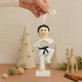 Load image into Gallery viewer, Decorative ornament of a martial artist in a white gi with a black belt, held by a hand against a neutral background.