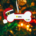 Load image into Gallery viewer, Personalized dog Christmas ornament featuring a bone shaped design with a glitter red Santa hat detail on one end, customizable name area centered on the ornament and customizable year displayed on the opposite side, smooth glossy finish with glitter accents, flat lightweight structure and red hanging ribbon, photographed on a snowy holiday styled background with pine branches, festive Christmas ornaments, dog themed props, and warm seasonal lighting for pet lovers.