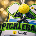 Load image into Gallery viewer, Personalized pickleball Christmas ornament featuring crossed pickleball paddles and a yellow pickleball on a shield style background, customizable name area on the front banner and customizable year area on the ball, glossy finish with raised 3D front details and flat 2D back, white hanging ribbon, displayed on a snowy pickleball court themed holiday background with pine branches and festive decorations.
