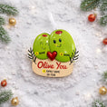 Load image into Gallery viewer, Decorative ornament with two smiling green olives holding a heart, surrounded by Christmas decorations on a snowy background.