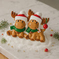 Load image into Gallery viewer, Two moose figurines wearing Santa hats and scarves on a snowy surface with Christmas decorations.