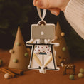 Load image into Gallery viewer, Decorative ornament of a person on a ladder with a camera, held by a hand against a festive background.