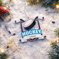 Load image into Gallery viewer, Personalized hockey Christmas ornament featuring a hockey puck and crossed hockey sticks design, customizable name area on the front banner and customizable year area on the puck, glossy finish with raised 3D front details and flat 2D back, white hanging ribbon, displayed on a snowy ice rink themed holiday background with hockey accessories and festive winter decorations.