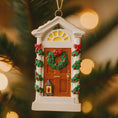Load image into Gallery viewer, Decorative Christmas ornament shaped like a house with a door and wreath, hanging against a blurred festive background.