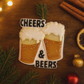 Load image into Gallery viewer, Personalized Cheers and Beers Christmas ornament featuring a raised 3D front with two frothy beer mugs clinking together. The design includes bold âCheersâ and âBeersâ lettering, detailed foam texture, and warm amber beer tones. The ornament has a flat 2D back for easy hanging. Hand-added personalization allows two names and a year. Styled on a rustic wooden background with Christmas lights, greenery, and festive decor for a cozy holiday atmosphere.