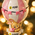 Load image into Gallery viewer, A personalized baby girl ornament featuring a cute bunny riding a pastel pink hot air balloon. Soft pink and white tones with delicate bow details create a sweet and cozy look. Handwritten personalization includes baby name and year. Mostly 2D ornament with raised 3D details on the front only, lightweight and easy to hang, perfect for baby first Christmas, nursery decor, and toddler keepsakes.