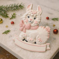 Load image into Gallery viewer, A glossy resin baby girl llama ornament features a white smiling llama with soft pink accents, raised textured fur details, pink bow and scarf elements, glitter highlights, and a smooth painted cartoon style finish, styled with warm holiday lights and nursery themed props, handwritten personalization showing the baby name on the front banner and the year placed on the llama body, designed as a mostly 2D ornament with raised 3D details on the front only.