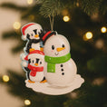 Load image into Gallery viewer, Christmas ornament with a snowman and two penguins on a tree with lights in the background