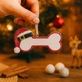 Load image into Gallery viewer, Personalized dog Christmas ornament featuring a bone shaped design with a glitter red Santa hat detail on one end, customizable name area centered on the ornament and customizable year displayed on the opposite side, smooth glossy finish with glitter accents, flat lightweight structure and red hanging ribbon, photographed on a snowy holiday styled background with pine branches, festive Christmas ornaments, dog themed props, and warm seasonal lighting for pet lovers.
