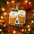 Load image into Gallery viewer, Personalized Cheers and Beers Christmas ornament featuring a raised 3D front with two frothy beer mugs clinking together. The design includes bold âCheersâ and âBeersâ lettering, detailed foam texture, and warm amber beer tones. The ornament has a flat 2D back for easy hanging. Hand-added personalization allows two names and a year. Styled on a rustic wooden background with Christmas lights, greenery, and festive decor for a cozy holiday atmosphere.