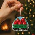Load image into Gallery viewer, Hand holding a Christmas ornament with red balls in a green wagon against a festive background.