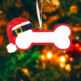 Load image into Gallery viewer, Personalized dog Christmas ornament featuring a bone shaped design with a glitter red Santa hat detail on one end, customizable name area centered on the ornament and customizable year displayed on the opposite side, smooth glossy finish with glitter accents, flat lightweight structure and red hanging ribbon, photographed on a snowy holiday styled background with pine branches, festive Christmas ornaments, dog themed props, and warm seasonal lighting for pet lovers.