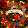 Load image into Gallery viewer, Personalized cardinal wreath ornament featuring a vibrant red cardinal bird surrounded by green holly leaves and red berries. Designed with a smooth glossy finish and a raised 3D front with a flat back for easy hanging. Includes free handwritten personalization with one name and year displayed on the banner at the top. Styled in a warm festive holiday setting with soft Christmas lights candy details and seasonal decor. A meaningful Christmas tree ornament symbolizing remembrance love and connection perfect