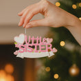 Load image into Gallery viewer, A personalized Little Sister ornament featuring soft pink layered lettering with heart accents and floral details. Designed in gentle pastel tones with a smooth resin finish and subtle glitter highlights. The ornament includes handwritten personalization with a baby name and year, created as a mostly 2D ornament with raised 3D details on the front only. Lightweight and easy to hang, perfect for celebrating a new baby sister and her first Christmas.