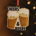 Load image into Gallery viewer, Personalized Cheers and Beers Christmas ornament featuring a raised 3D front with two frothy beer mugs clinking together. The design includes bold âCheersâ and âBeersâ lettering, detailed foam texture, and warm amber beer tones. The ornament has a flat 2D back for easy hanging. Hand-added personalization allows two names and a year. Styled on a rustic wooden background with Christmas lights, greenery, and festive decor for a cozy holiday atmosphere.